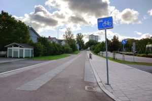 Valokuvassa kapea tyhjä katu jonka ajorata punertavaa asfalttia, keskellä ja reunoilla pitkittäiset laattakaistaleet. Oikealla laatoitettu jalkakäytävä jonka reunassa etualalla tolpassa pyöräkadun liikennemerkki.
